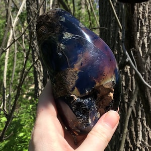 May include: A large, dark purple and black gemstone with a rough, irregular surface. The gemstone has a shiny, iridescent finish and is held in a hand.