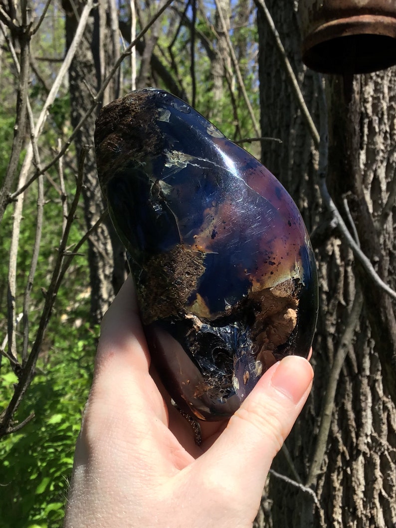 May include: A large, dark purple and black gemstone with a rough, irregular surface. The gemstone has a shiny, iridescent finish and is held in a hand.