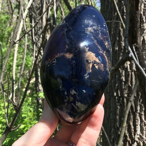 May include: A black obsidian egg with white and brown inclusions. The egg is smooth and polished, and it is being held in a hand.