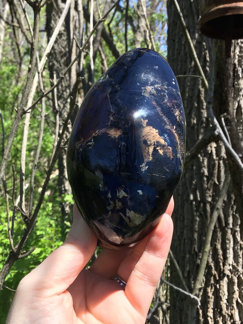 May include: A black obsidian egg with white and brown inclusions. The egg is smooth and polished, and it is being held in a hand.