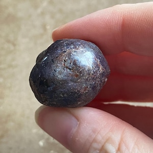 May include: A dark purple, rounded gemstone with a glossy surface. The stone has a slightly irregular shape and appears to be held between fingers. The gem's color varies from deep purple to nearly black, with some lighter, reflective areas.
