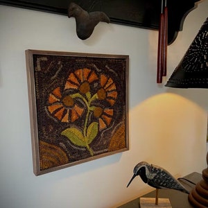 May include: A framed needlepoint artwork featuring a stylized floral design with orange, brown, and green colors. The artwork is displayed on a white wall.