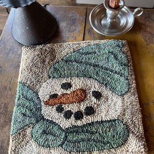 May include: A hand-stitched rug featuring a whimsical snowman with a green hat and a brown carrot nose. The rug is made with a variety of textures and colors, creating a unique and charming design.
