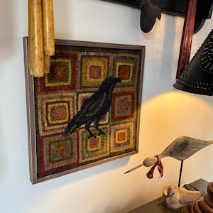May include: A framed, square, folk art style textile art piece featuring a black crow. The artwork is set against a patchwork background of squares in shades of green, red, and gold. The frame is a dark brown color.