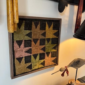 May include: A framed needlepoint wall hanging with a geometric pattern of brown, green, and pink stars on a black background.