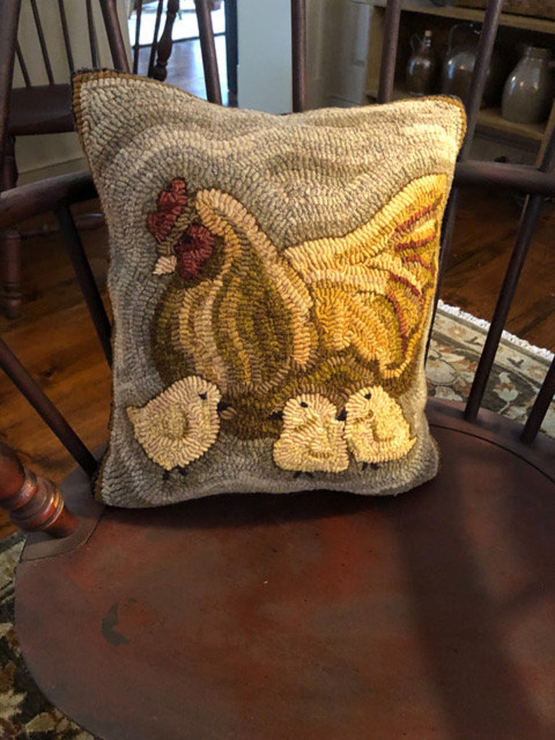 Puede incluir: Una almohada gris y marr&oacute;n con un dise&ntilde;o bordado de una gallina amarilla y tres pollitos. La almohada est&aacute; sentada en una silla de madera.