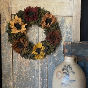 May include: A fall-themed wreath made of felt with sunflowers, brown flowers, and green leaves. The wreath is hanging on a rustic wooden door.