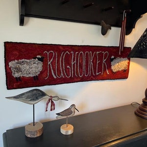 May include: A red rug with the text "RUGHOOKER" and two sheep. Below, a wooden bird sculpture with a red ribbon and a smaller bird sculpture. A black lamp with a perforated shade sits on a black dresser.