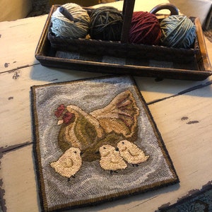 Puede incluir: Una alfombra hecha a mano que representa una gallina marr&oacute;n y blanca con tres pollitos amarillos. La alfombra est&aacute; sobre una mesa de madera blanca con una cesta de lana en el fondo.