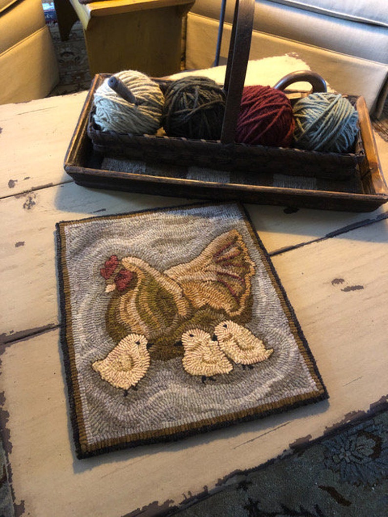 Puede incluir: Una alfombra hecha a mano que representa una gallina marr&oacute;n y blanca con tres pollitos amarillos. La alfombra est&aacute; sobre una mesa de madera blanca con una cesta de lana en el fondo.