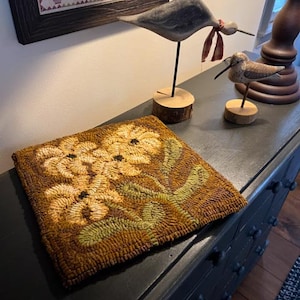 May include: A square, hand-stitched rug with a floral design in shades of yellow, green, and brown. The rug is placed on a black dresser, with wooden bird sculptures and a framed artwork in the background.