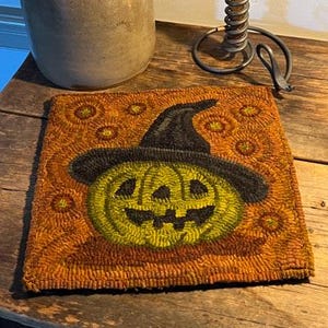May include: A brown and orange rug with a black and green pumpkin wearing a black witch hat. The pumpkin has a carved jack-o'-lantern face.