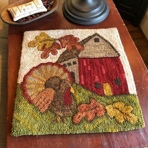 May include: A hand-stitched wool rug depicting a red barn with a turkey in front of it. The barn is surrounded by green grass and fall leaves. The rug is perfect for adding a touch of autumn charm to your home.