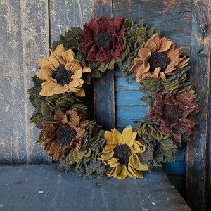 May include: A felt sunflower wreath with a rustic wooden background. The wreath is made of brown, yellow, and green felt and features a variety of sunflower designs.