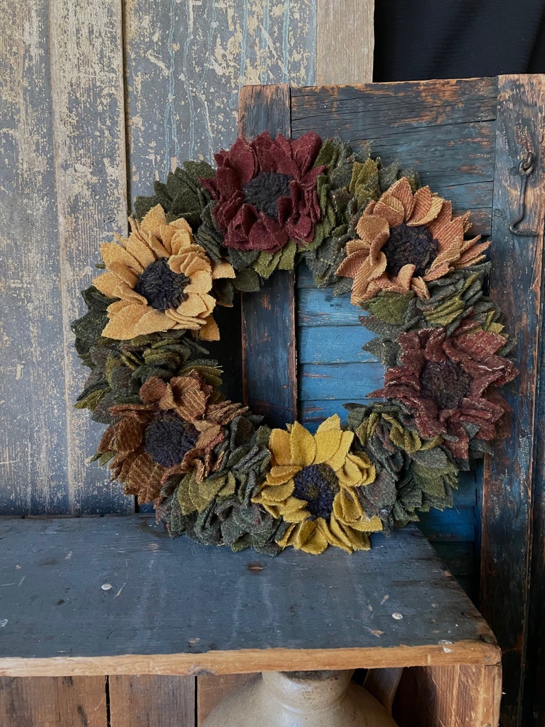 May include: A felt sunflower wreath with a rustic wooden background. The wreath is made of brown, yellow, and green felt and features a variety of sunflower designs.