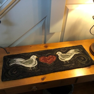 May include: A brown wooden table with a black and green rug featuring two white birds and a red heart. A black candle holder with a red candle is on the left side of the table. A black lamp with a brown base is on the right side of the table.