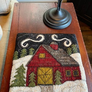 May include: A rectangular, hand-stitched rug depicting a winter scene. A red cabin with a yellow door and windows is surrounded by snow, green trees, and a dark night sky with white swirls. The rug is placed on a wooden surface.