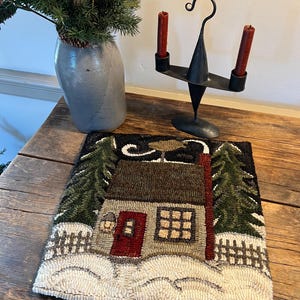 May include: A rustic, hand-hooked rug depicting a winter cabin scene with snow, trees, and a red door. The rug is placed on a weathered wooden table, alongside a gray ceramic vase with greenery and a black metal candle holder with red candles.