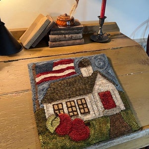 May include: A hand-stitched wool rug depicting a white schoolhouse with a red door, a waving American flag, and two red apples in the foreground. The rug is displayed on a wooden desk with books and a quill pen.