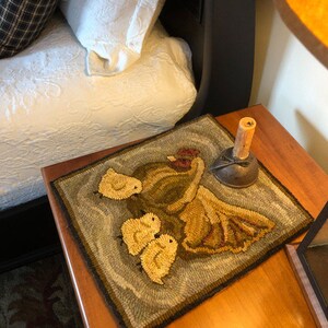 Puede incluir: Una alfombra hecha a mano con un esquema de color marr&oacute;n, verde y amarillo. La alfombra presenta un dise&ntilde;o de una madre p&aacute;jaro y tres polluelos. La alfombra est&aacute; sobre una mesa de madera con una l&aacute;mpara y un objeto decorativo.