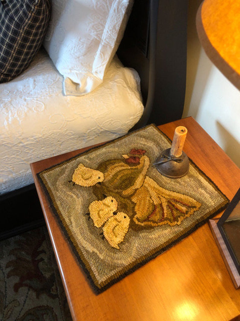 Puede incluir: Una alfombra hecha a mano con un esquema de color marr&oacute;n, verde y amarillo. La alfombra presenta un dise&ntilde;o de una madre p&aacute;jaro y tres polluelos. La alfombra est&aacute; sobre una mesa de madera con una l&aacute;mpara y un objeto decorativo.