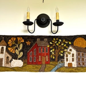 May include: A hand-stitched wool rug depicting a quaint village scene with a church, a red house, a white house, a tree, a sheep, and a full moon. The rug is hung on a wall with a black candle sconce above it.
