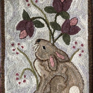 May include: Hand-hooked rug featuring a brown rabbit with pink ears and a white tail, set against a backdrop of flowers in shades of purple and green. The rug has a textured, off-white background and a brown border.