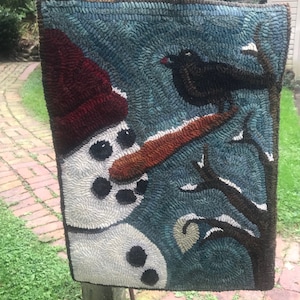 May include: A hand-stitched wool rug depicting a snowman with a red hat and a black bird perched on a branch. The background is a blue-green color.