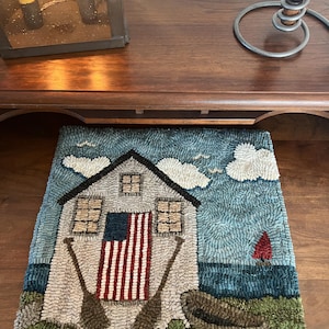 May include: Hand-hooked rug featuring a coastal scene with a house, American flag, and oars. The rug has a blue sky with clouds, a red sailboat, and a green grassy area. The rug is rectangular.