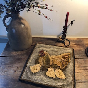 Puede incluir: Una alfombra hecha a mano que representa una gallina marr&oacute;n y amarilla con tres pollitos amarillos. La alfombra tiene un fondo gris y un borde negro.