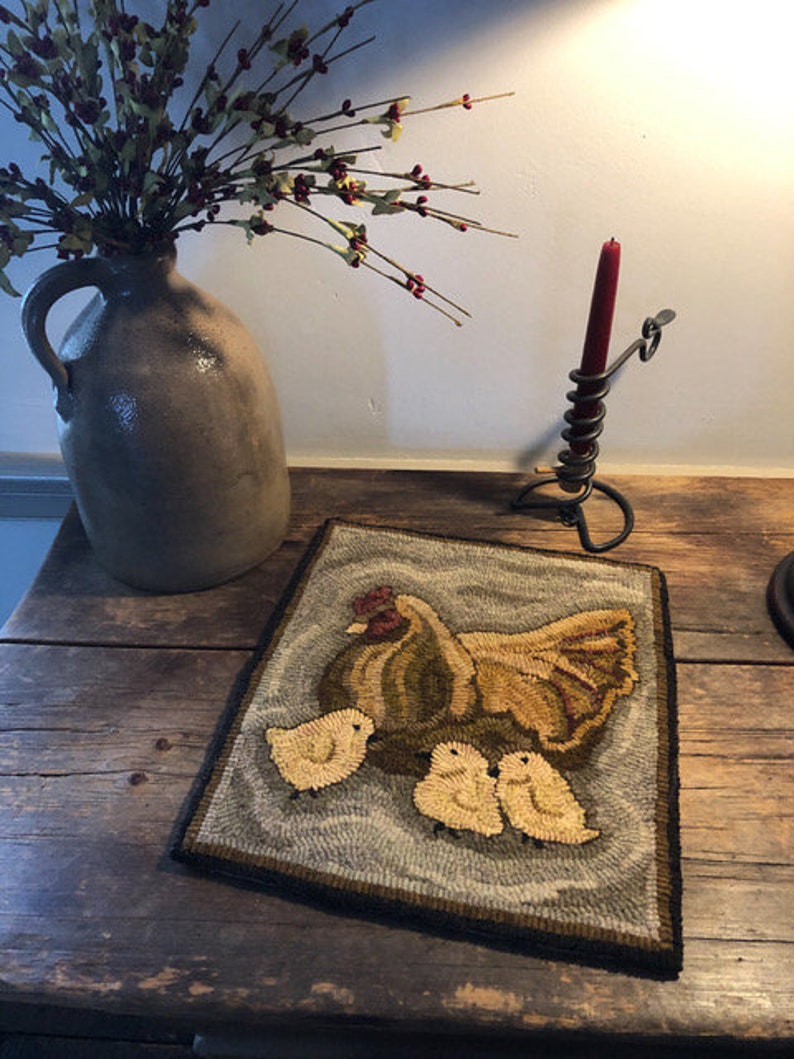 Puede incluir: Una alfombra hecha a mano que representa una gallina marr&oacute;n y amarilla con tres pollitos amarillos. La alfombra tiene un fondo gris y un borde negro.