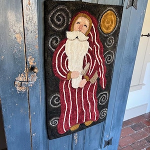 May include: A hand-stitched wool rug depicting a whimsical Santa Claus figure in a red and white striped robe, holding a sack. The background features a dark blue sky with swirling clouds and a yellow moon.