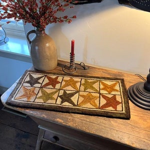 May include: A brown wooden table with a drawer and a brown and beige needlepoint runner with a star pattern. The runner is on top of the table. There is a red candle on a metal candlestick and a brown ceramic pitcher with dried flowers on the table.