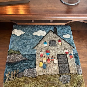 May include: Hand-hooked rug featuring a coastal scene with a gray house, blue water, and white clouds. The house has a dark gray roof and colorful hanging lanterns. The rug is made of textured yarn and has a rectangular shape.