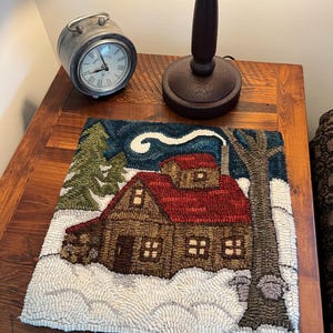 May include: A hooked rug showing a winter cabin scene with a red roof and brown walls. The rug features a snowy landscape, a green fir tree, and a bare tree. A vintage clock and a lamp are on the wooden table, adding to the rustic charm.