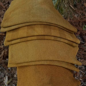 May include: A stack of four mustard-colored, textured fabric pillows. The pillows are folded and layered, with visible seams and varying textures. The background features a tree trunk and brown foliage.