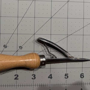 May include: A metal sewing awl with a light brown wooden handle, positioned on a cutting mat with inch markings. The tool features a pointed metal needle and a hinged metal lever mechanism attached to the handle, measuring 4.5 inches in length.