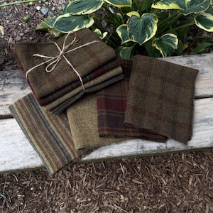 May include: A collection of folded fabric swatches in various earth tones including olive green, brown, and deep red. The textiles feature diverse patterns such as subtle plaid, windowpane checks, and vertical stripes, with one stack tied together by twine.