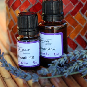 May include: Two small brown glass bottles with black caps and purple labels that say "Lavender Essential Oil" and "Intermedia" with the volume in milliliters. The bottles are sitting on a woven bamboo surface with a sprig of lavender flowers.