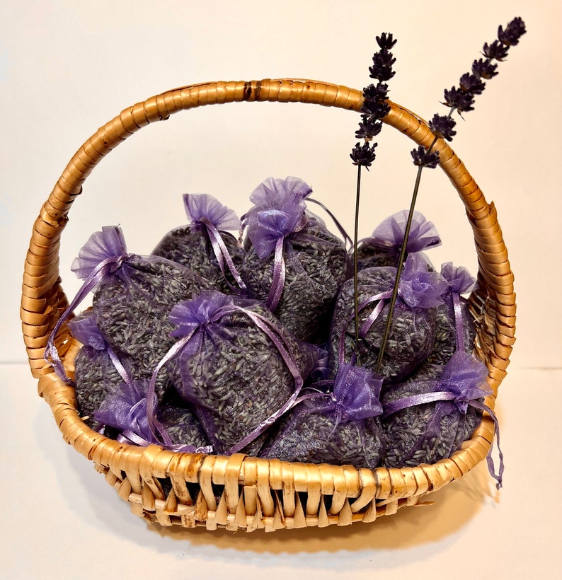 May include: A wicker basket filled with lavender sachets. The sachets are made of purple tulle and are tied with purple ribbon. The basket is decorated with sprigs of lavender.