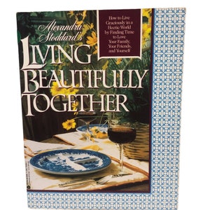 Pode incluir: Capa do livro "Living Beautifully Together" de Alexandra Stoddard. A imagem apresenta uma mesa posta com pratos azuis e brancos, taças de vinho e flores amarelas. O subtítulo é: "Como viver com graça num mundo agitado, encontrando tempo para amar a sua família, amigos e a si mesmo."