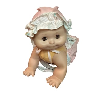 May include: A vintage baby doll wearing a pink bonnet and a yellow outfit. The doll is in a crawling position, with its arms and legs outstretched. The doll has brown eyes and a soft expression. The bonnet features a striped design.