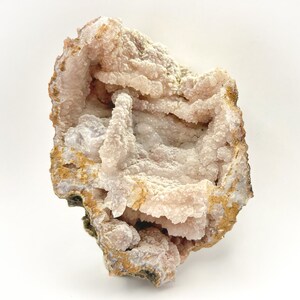 May include: A geode specimen with a rough, textured exterior in shades of tan, brown, and cream. The interior reveals a cluster of pale pink crystal formations. The natural stone is displayed against a white background.