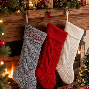 Op de afbeelding: Drie gebreide kerstsokken hangen op een rij. De eerste is grijs met het woord Dad geborduurd in rode draad. De tweede is effen rood en de derde is effen wit. Elke sok heeft een kabelbreipatroon en een boord aan de bovenkant.