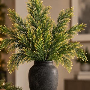 May include: A dark charcoal gray ceramic vase with a matte, textured finish holds a dense arrangement of artificial evergreen branches. The branches feature deep green needles tipped with small, bright yellow buds throughout the foliage.