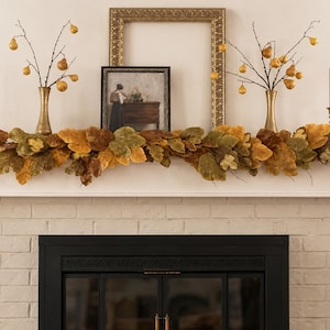 May include: A fireplace mantel decorated with a thick garland of autumn-coloured leaves in shades of brown, gold, and green. Two brass vases hold branches with small yellow pears, flanked by two tall, thin, light-coloured taper candles in brass holders.