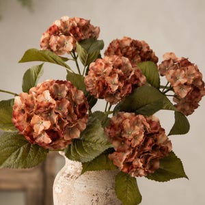 May include: A bouquet of artificial hydrangea flowers in shades of rust, copper, and muted orange. The round flower heads feature dense clusters of petals with green veining, accompanied by large, serrated green leaves, all arranged in a textured vase.
