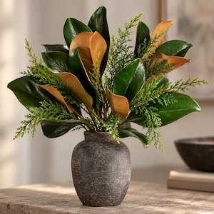 Peut inclure: Un vase en céramique gris foncé, texturé et arrondi, contenant un arrangement de feuilles de magnolia artificielles et de brins de cèdre verts. Les feuilles de magnolia présentent des dessus vert foncé brillants et des dessous brun clair, entremêlés de fines branches de cèdre.