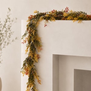 May include: A decorative garland featuring a dense arrangement of faux foliage in shades of green, yellow, and orange. The flexible vine-like structure is draped over a white surface, showcasing small, pointed leaves that create a layered texture.