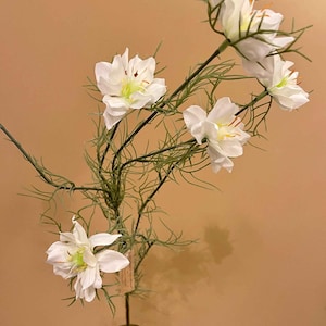 May include: A floral display of white flowers with yellow centres and green detailing. The flowers are arranged on thin green stems with delicate leaves, presented in a green glass vase. The background is a plain tan colour.